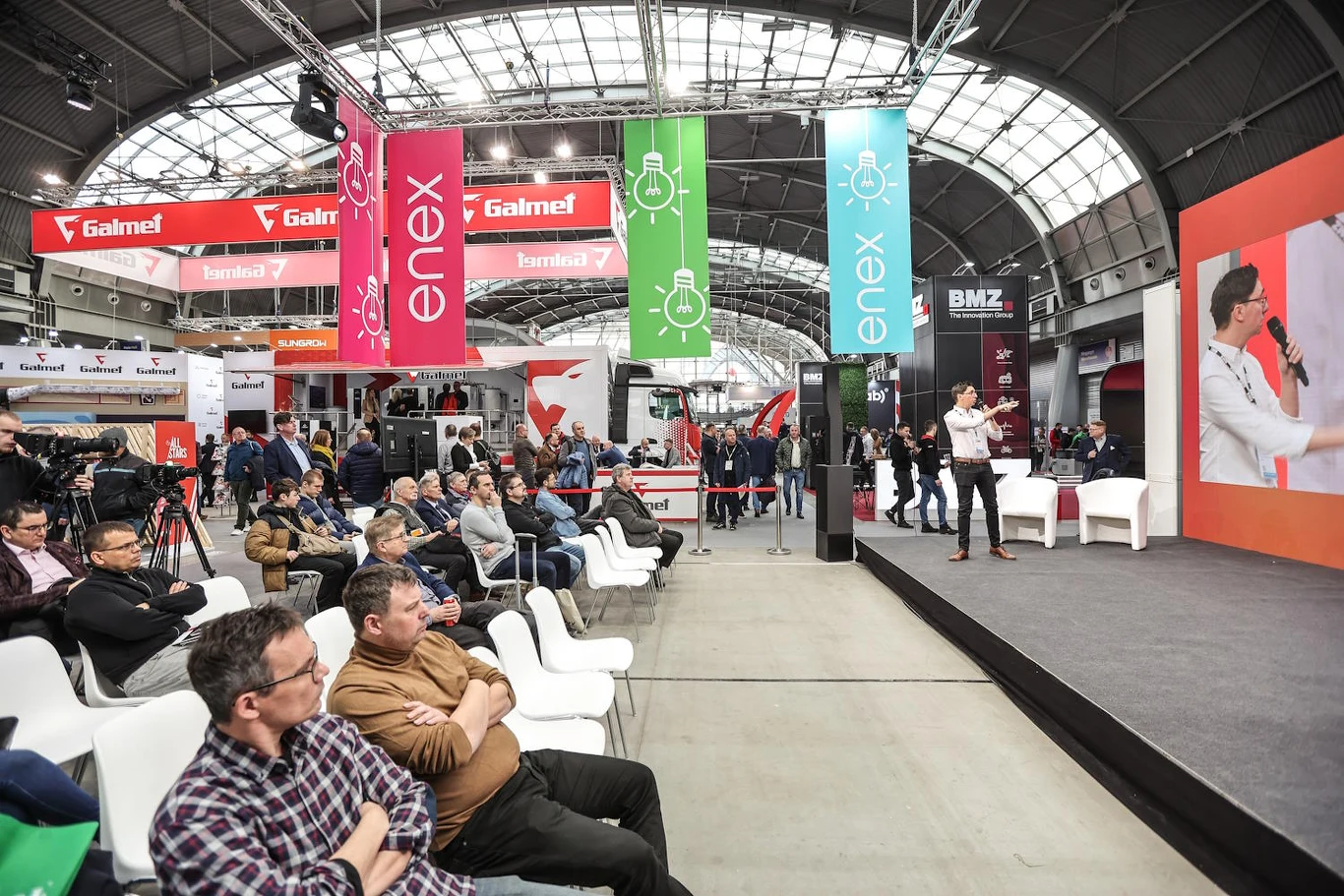 Publiczność uważnie słucha osoby przemawiającej na scenie podczas targów lub konferencji odbywających się w nowoczesnej hali wystawienniczej. W tle widoczne są kolorowe banery reklamowe oraz stanowiska wystawców.
