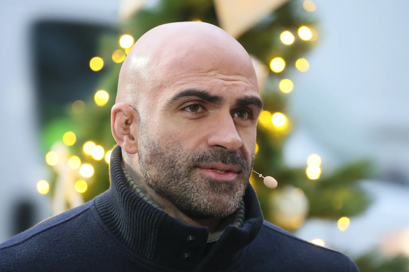 Łysy mężczyzna z mikrofonem na twarzy, ubrany w ciemny golf, stoi na tle udekorowanego świątecznymi światełkami drzewka.