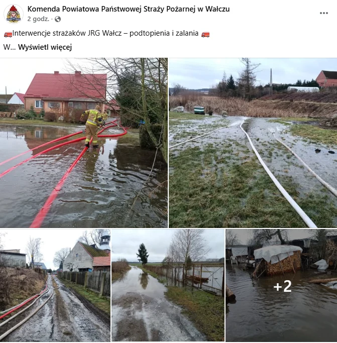 Strażak w żółtym kombinezonie rozkłada węże strażackie na podwórku zalanym wodą, wokół widoczne są podtopione domy i zabudowania gospodarcze, a w tle rozciągają się mokre pola i ogródki otoczone wodą.