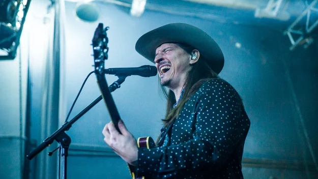 Muzyk w kapeluszu śpiewa do mikrofonu, trzymając gitarę elektryczną na scenie oświetlonej niebieskim światłem, z intensywną ekspresją twarzy.