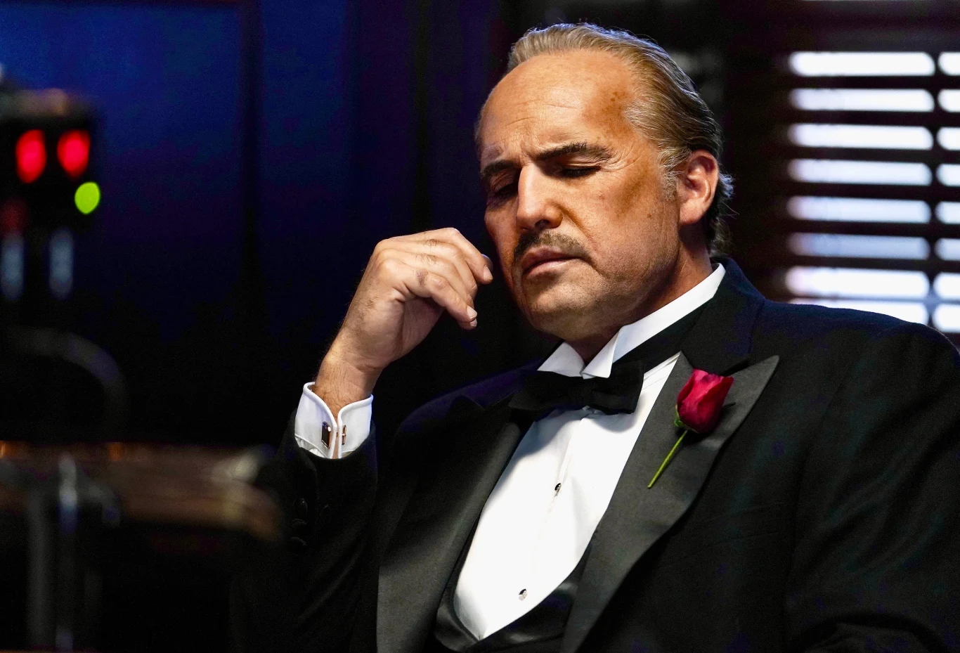 Billy Zane jako Marlon Brando Mężczyzna w smokingu z czerwoną różą w butonierce siedzi z zamyśloną miną w ciemnym, eleganckim wnętrzu, światło wpada przez żaluzje, podkreślając jego twarz.