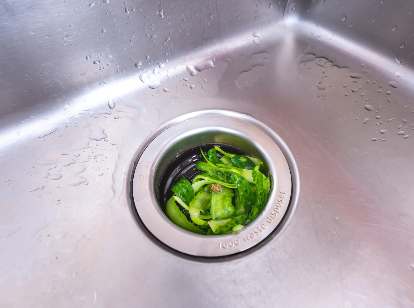 Czy stosowanie młynków do mielenia resztek jest legalne? Zielone obierki i resztki warzyw znajdują się w odpływie zlewu kuchennego ze śladami wody wokół, stal nierdzewna dominuje w otoczeniu.