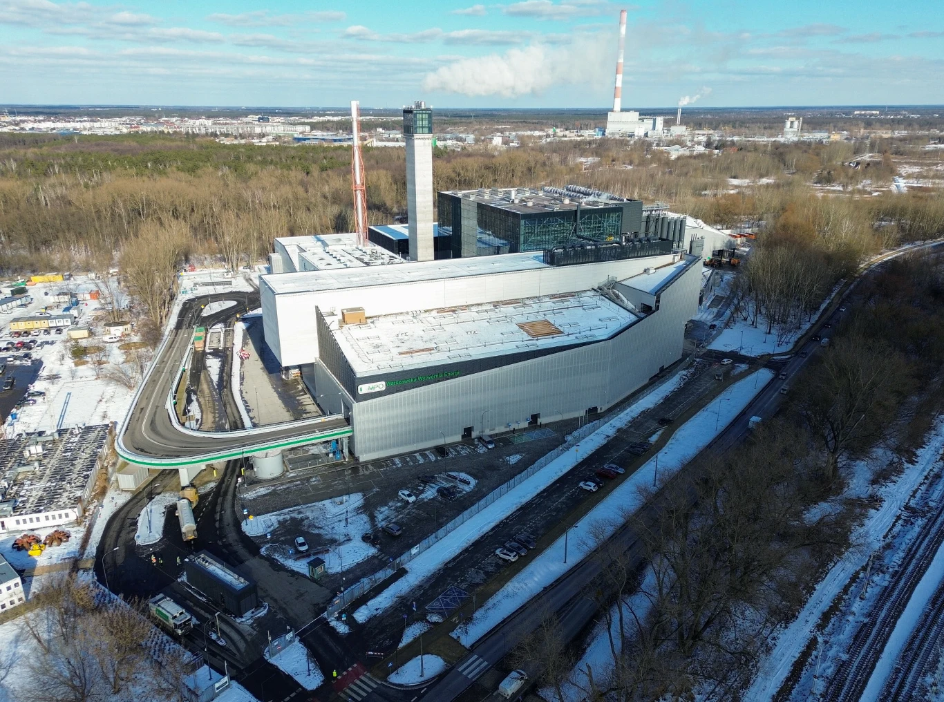 Nowoczesna spalarnia śmieci z dużymi przemysłowymi budynkami, kominami oraz przyległymi infrastrukturami, otoczona lasem i pokrytym śniegiem terenem. Po lewej stronie widoczne drogi i obiekty technologiczne, a w tle większy kompleks przemysłowy.
