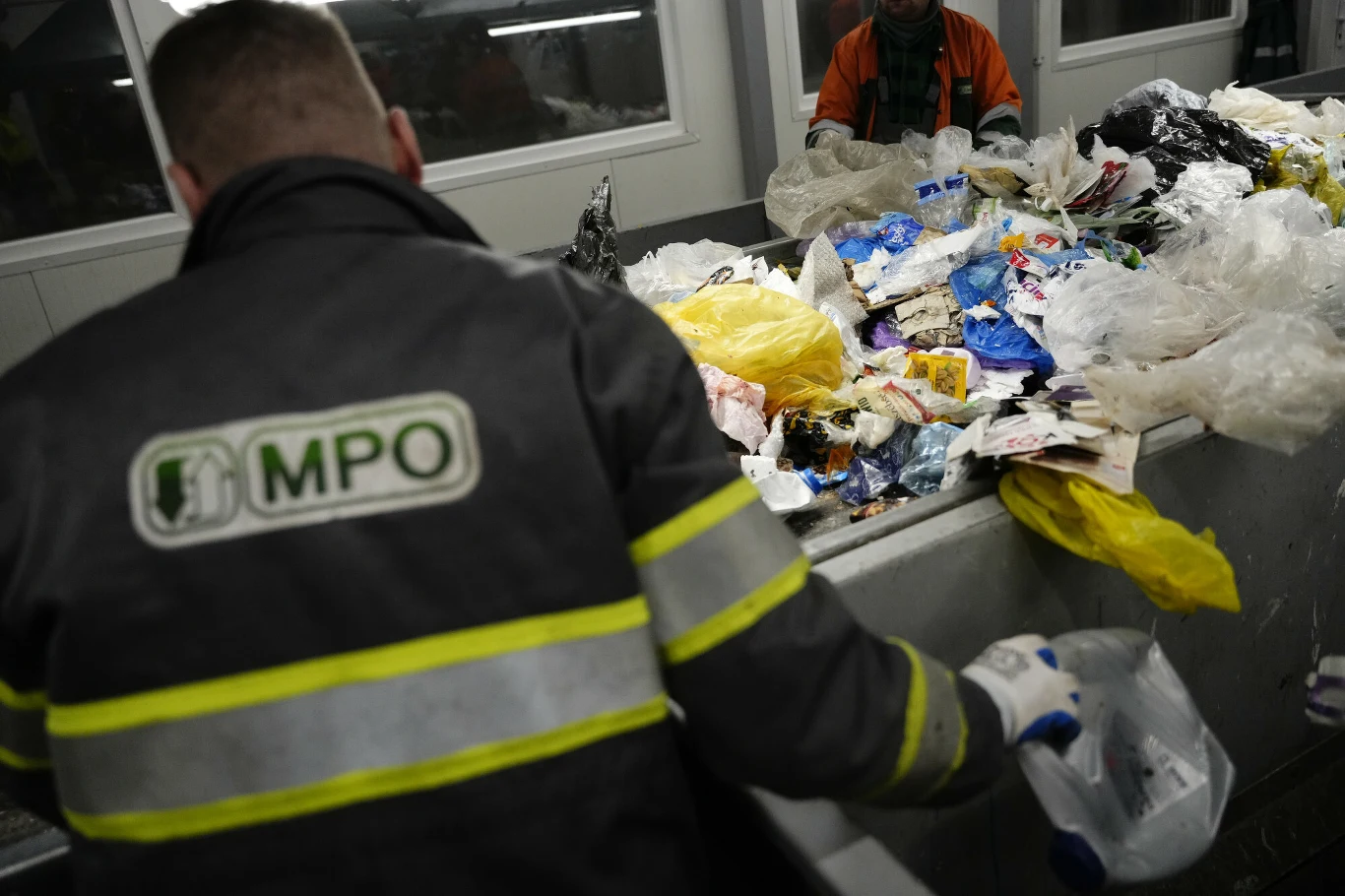 Pracownicy w odblaskowych kurtkach sortują różnorodne odpady, głównie plastikowe worki, butelki i inne śmieci, na taśmie sortowniczej w zakładzie przetwarzania odpadów.