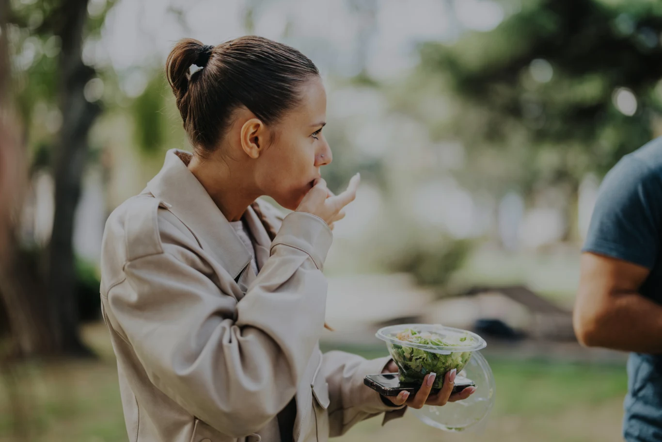 Kobieta w jasnym płaszczu je sałatkę na świeżym powietrzu, liżąc palce trzymając przezroczyste opakowanie z zieloną sałatą. W tle rozmyte drzewa i druga osoba częściowo widoczna po prawej stronie.