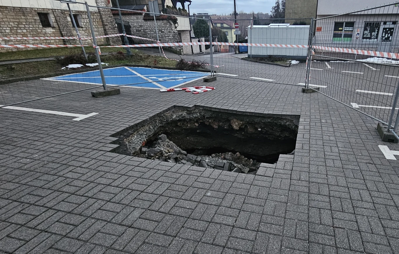 Duża zapadlina w nawierzchni parkingu otoczona taśmą ostrzegawczą i metalowym ogrodzeniem, w tle widoczne miejsca parkingowe oraz fragment niebieskiego stanowiska dla osób niepełnosprawnych.