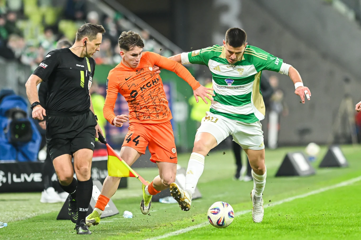 Piłkarz w zielono-białym stroju rywalizuje o piłkę z zawodnikiem w pomarańczowym stroju przy linii bocznej boiska, a sytuacji przygląda się sędzia z gwizdkiem.