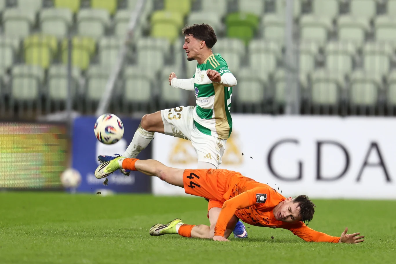 Dwóch piłkarzy rywalizujących o piłkę podczas meczu, jeden z nich w pomarańczowym stroju leży na murawie, drugi w biało-zielonym stroju unosi się w powietrzu po kontakcie fizycznym, w tle pusty stadion.