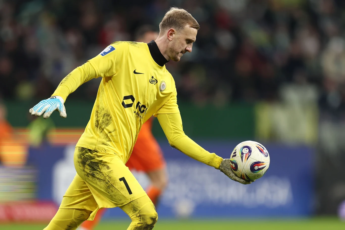 Bramkarz w żółtym stroju przygotowujący się do wyrzutu piłki podczas meczu piłki nożnej, trzyma piłkę obiema rękami, a jego odzież jest ubrudzona w okolicach kolan.