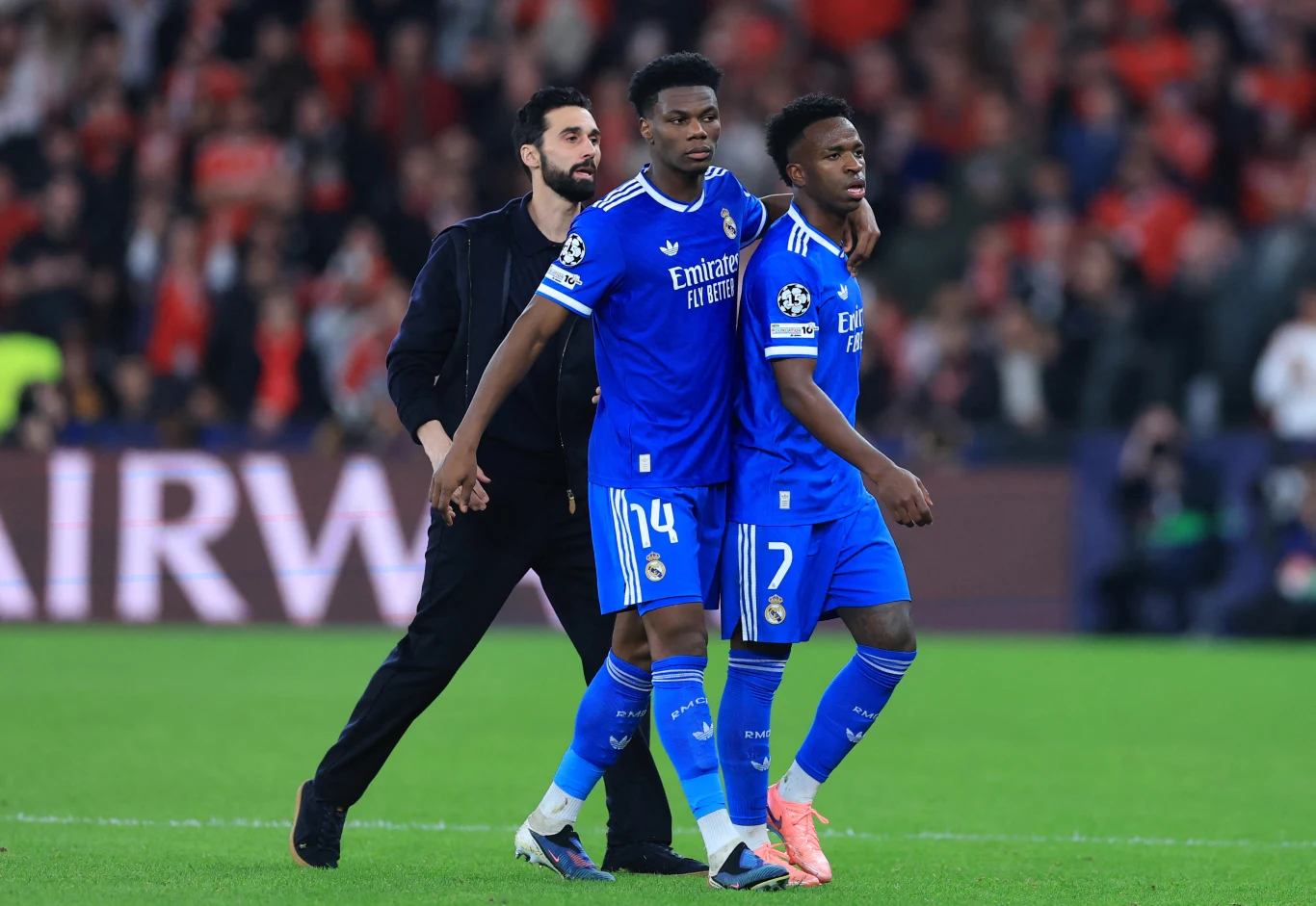 Dwóch piłkarzy w niebieskich strojach drużyny Real Madryt idzie razem po murawie, jeden z nich jest wspierany przez kolegę z zespołu, w tle widoczne są trybuny z kibicami.