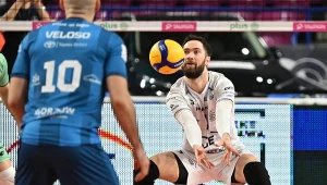 Zawodnik drużyny siatkarskiej w białym stroju odbija piłkę sposobem dolnym podczas meczu, w tle widoczny zawodnik drużyny przeciwnej w niebieskim stroju z numerem 10.