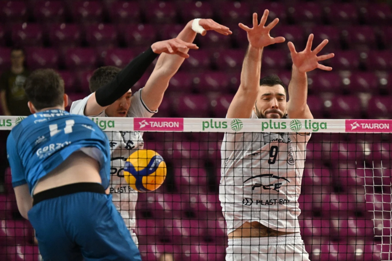 Dwóch zawodników w białych koszulkach blokuje piłkę siatkową przy siatce, podczas gdy trzeci zawodnik w niebieskim stroju wykonuje atak. W tle widać puste, czerwone trybuny oraz bandy reklamowe PlusLiga i Tauron.