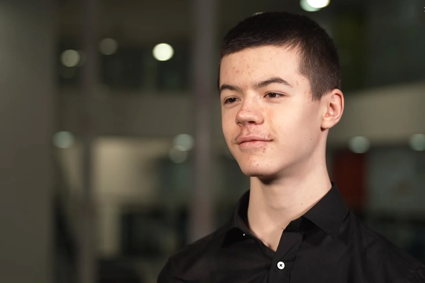 Młody mężczyzna o krótkich ciemnych włosach, ubrany w czarną koszulę, stoi na tle rozmytego, nowoczesnego wnętrza z jasnym oświetleniem.