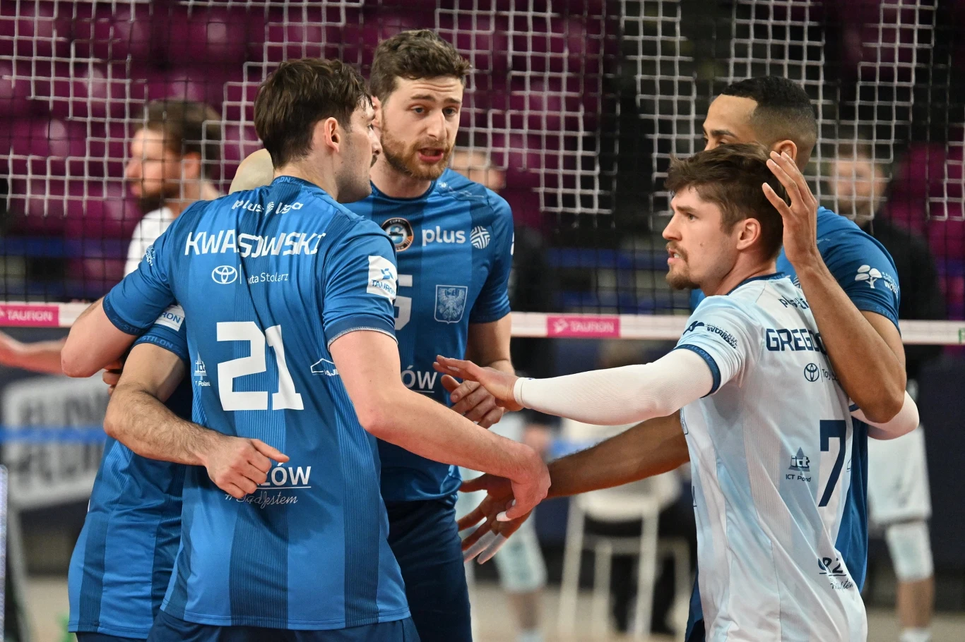 Grupa siatkarzy w sportowych strojach na boisku, zawodnicy jednej drużyny ubrani w niebieskie koszulki i szorty, skupieni wokół siebie podczas meczu, po drugiej stronie siatki zawodnik drużyny w białej koszulce, widoczne sportowe emocje i interakcje mi...