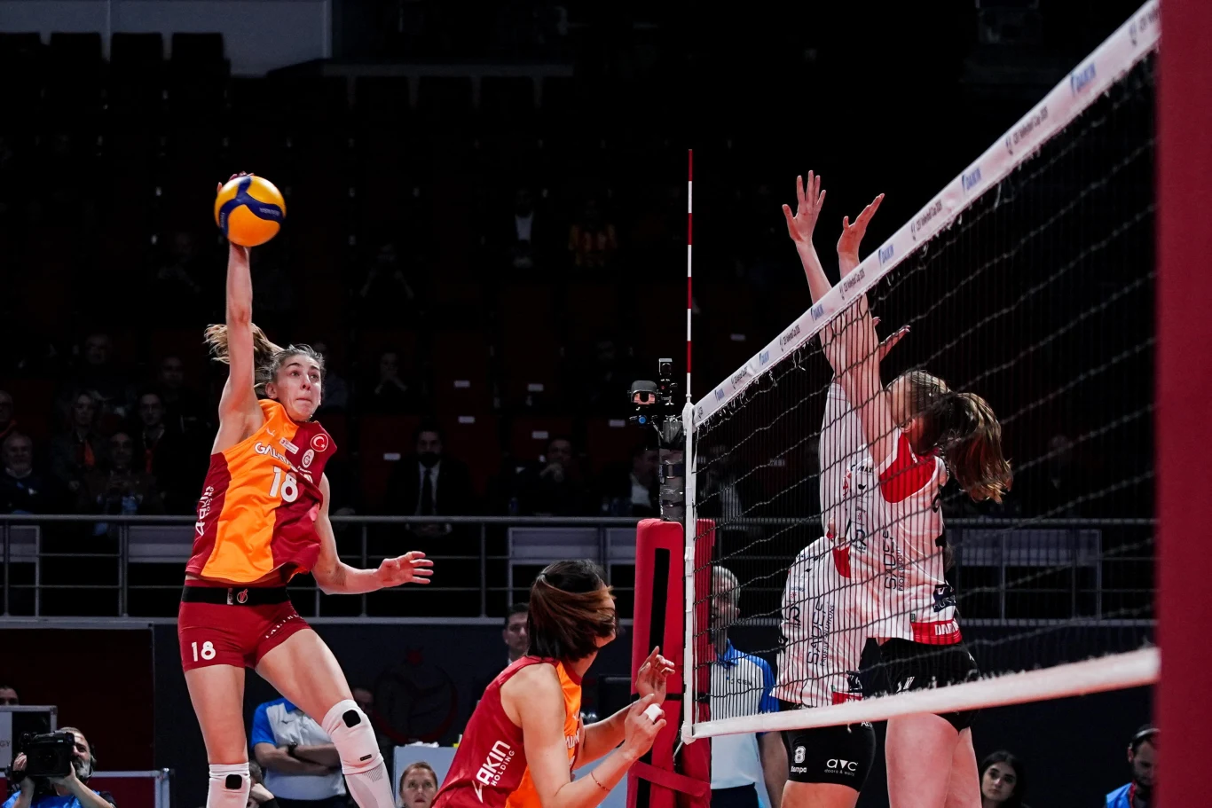 Zawodniczka w pomarańczowo-czerwonym stroju wykonuje atak na siatce podczas meczu siatkówki, druga drużyna po drugiej stronie siatki próbuje zablokować piłkę. W tle widoczna publiczność na trybunach.