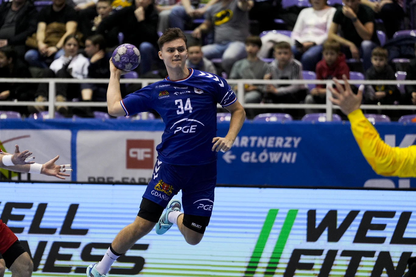 Zawodnik w niebieskim stroju z numerem 34 wykonuje dynamiczny rzut piłką ręczną podczas meczu, w tle widoczni kibice oraz inni zawodnicy.
