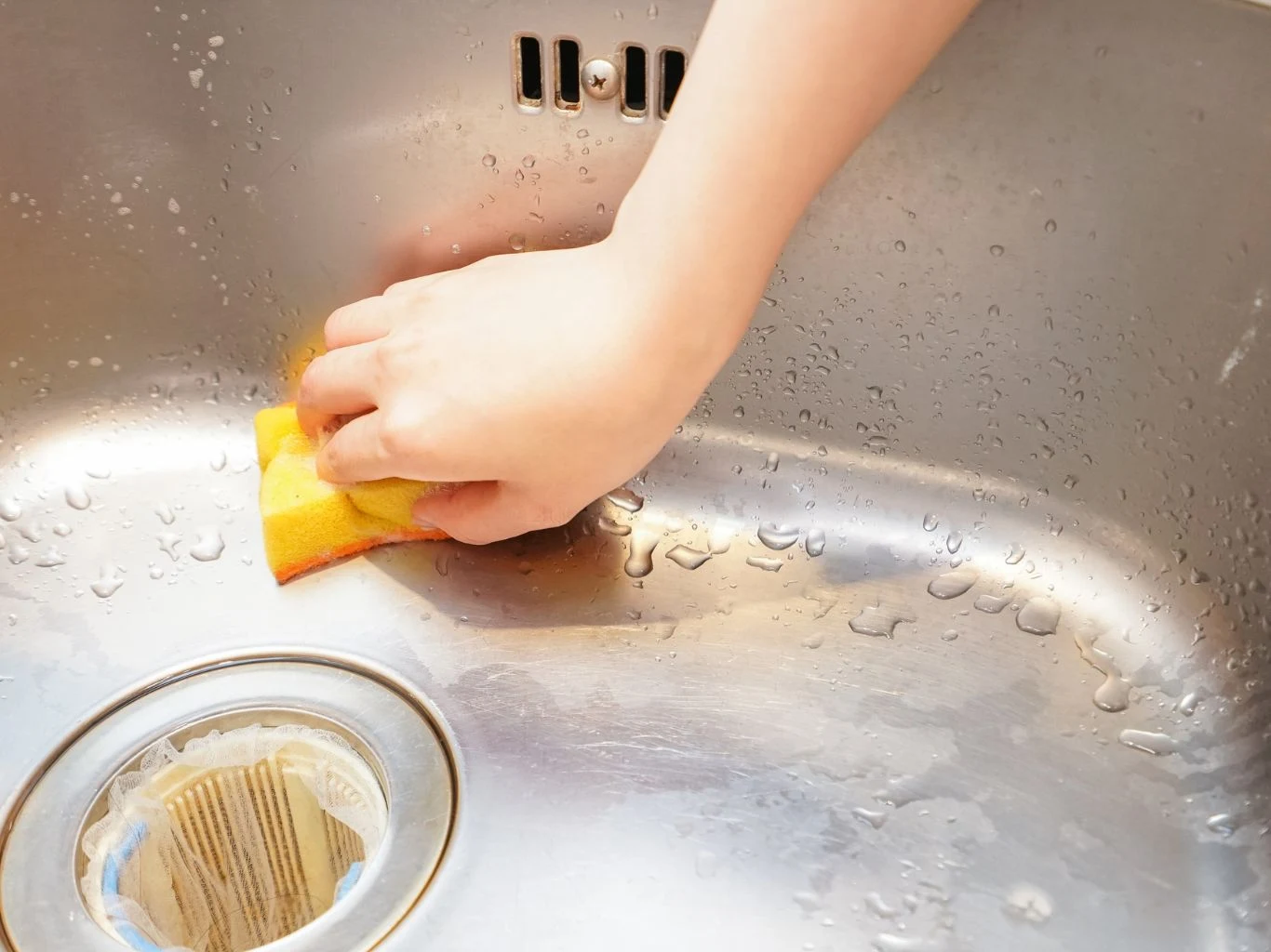 Gąbka luffa przyda się również do sprżatania w domu. Dłoń trzymająca żółtą gąbkę czyści metalowy zlew kuchenny, z widocznymi kroplami wody wokół odpływu.
