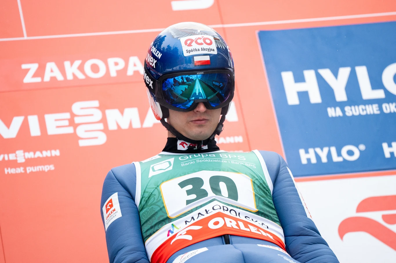 Skoczek narciarski w kombinezonie startowym i kasku z okularami ochronnymi stoi na tle banerów sponsorskich podczas zawodów.