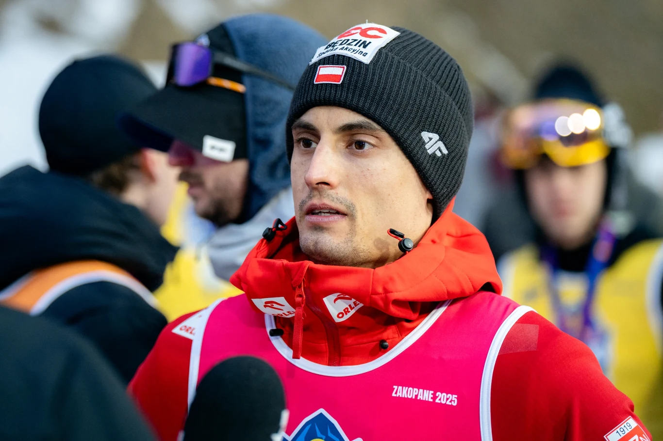 Sportowiec w czapce zimowej i czerwonym stroju, udzielający wywiadu po zawodach, na tle innych ludzi w odzieży sportowej na śnieżnym terenie.