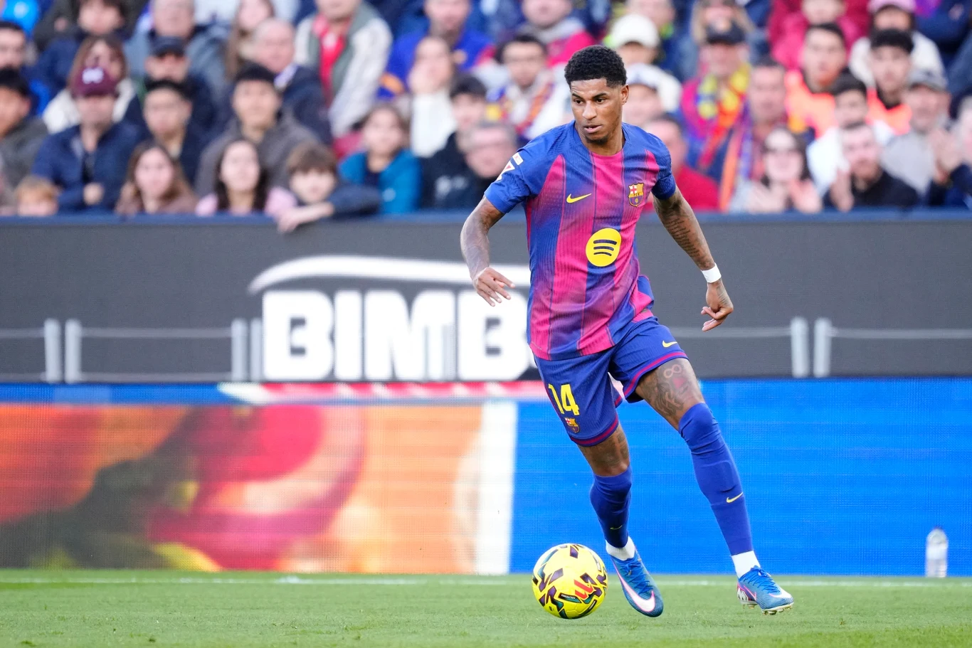 Piłkarz w niebiesko-bordowej koszulce z numerem 14 prowadzi piłkę po murawie stadionu, w tle widoczna publiczność i banery reklamowe.