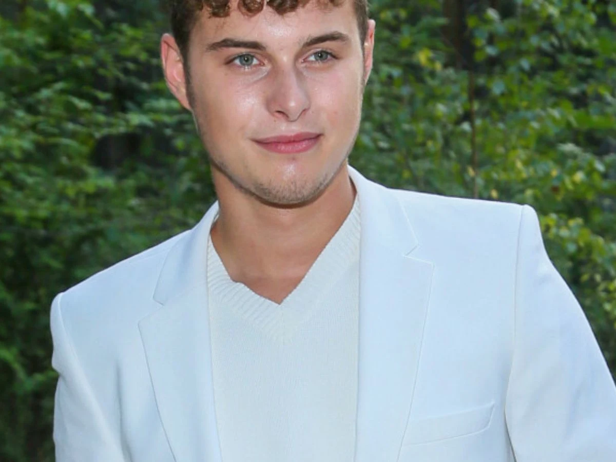 Adam Zdrójkowski Młody mężczyzna z krótkimi kręconymi włosami ubrany w elegancką białą marynarkę oraz biały sweter, stojący na tle zieleni lasu.