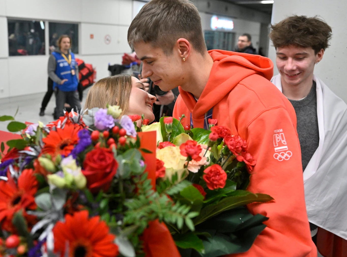 Powitanie Władimira Semirunnija na Okęciu Młody mężczyzna w czerwonej bluzie z logo Polskiego Komitetu Olimpijskiego uśmiecha się, przytulając kobietę z dużym bukietem kolorowych kwiatów, w tle widać jeszcze jednego uśmiechniętego młodego mężczyznę.