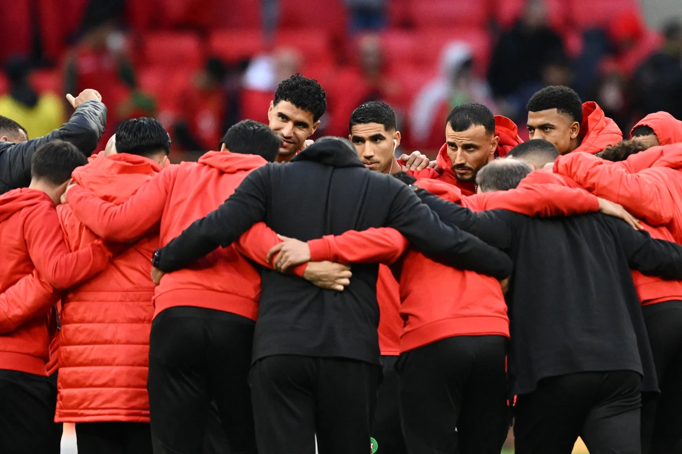 Romain Saiss (na fotografii ustawiony przodem jako drugi od prawej Grupa sportowców ubrana w czerwono-czarne stroje stoi w zwartym kręgu na stadionie, obejmując się nawzajem, w tle trybuny.