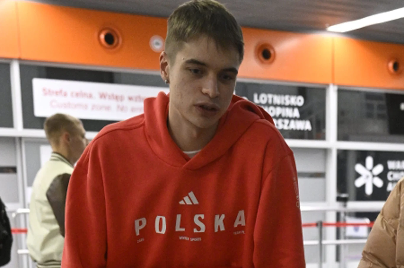 Władimir Semirunnij na lotnisku Okęcie w czerwonej bluzie z napisem 'Polska', w tle informacyjne szyldy i pasażerowie.
