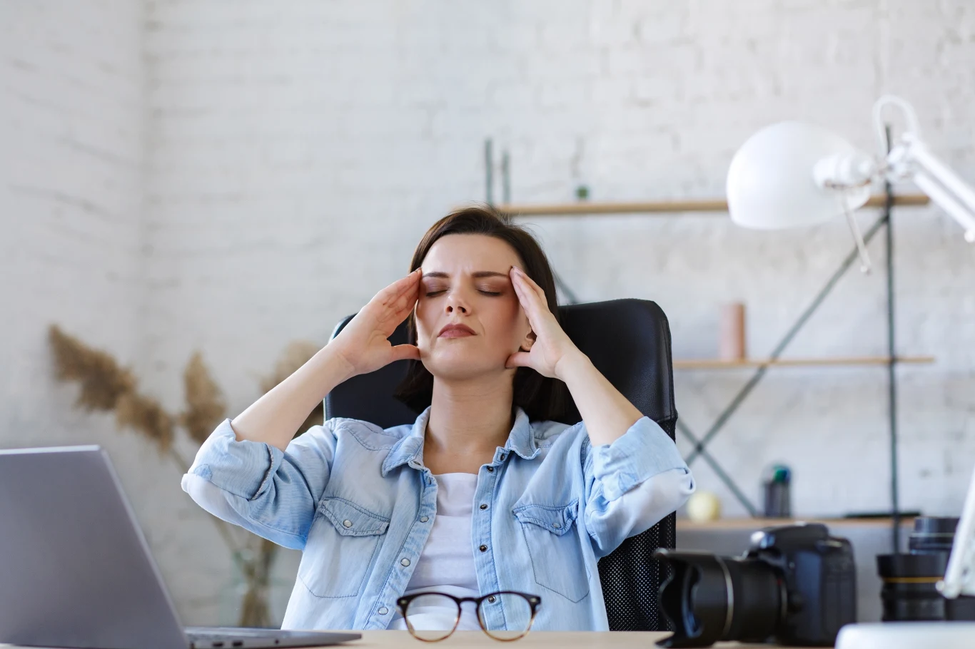 Zdaniem niektórych męczy nie tyle rodzicielstwo, co łącznie go z innymi obowiązkami Kobieta w jeansowej koszuli siedzi przy biurku, masuje skronie zamkniętymi oczami, sprawiając wrażenie zmęczonej lub zestresowanej. Na biurku przed nią stoi laptop i okulary, w tle widać białą lampkę oraz rozmyte elementy wyposażenia biura.