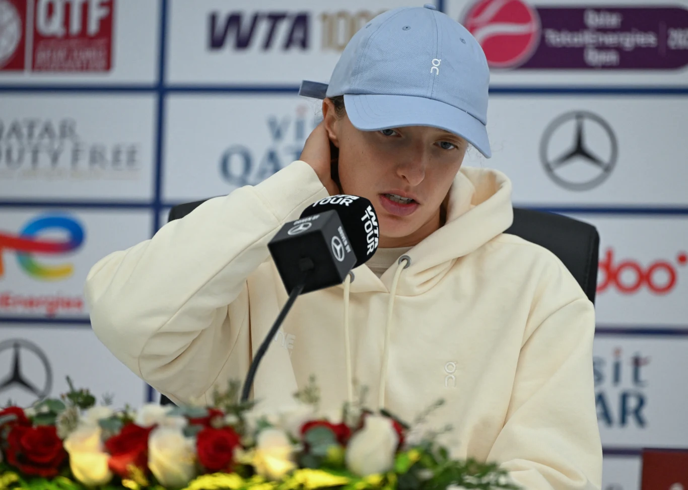 Sportowiec w jasnej bluzie i błękitnej czapce z daszkiem udziela wywiadu podczas konferencji prasowej, siedząc przy mikrofonie z nazwą turnieju, na stole leży bukiet kwiatów, w tle widoczne logotypy sponsorów i organizatorów turnieju tenisowego.