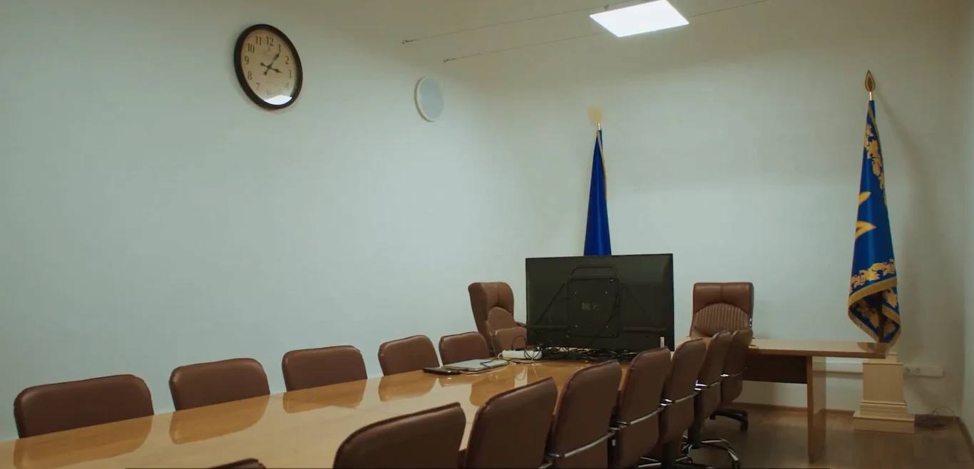 Sala konferencyjna z długim stołem otoczonym krzesłami, dużym monitorem na jednym z końców stołu, flagą umieszczoną w rogu oraz zegarem ściennym zawieszonym nad miejscem spotkań. Jasne ściany oraz sufit z oświetleniem LED podkreślają formalny i oficjal...