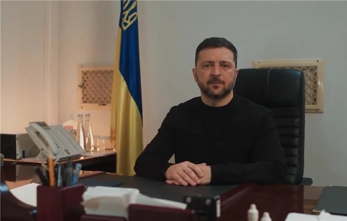 Mężczyzna w czarnym swetrze siedzi za biurkiem w gabinecie, za nim stoi flaga w niebiesko-żółtych barwach. Na biurku znajduje się telefon, kilka butelek z wodą oraz dokumenty.