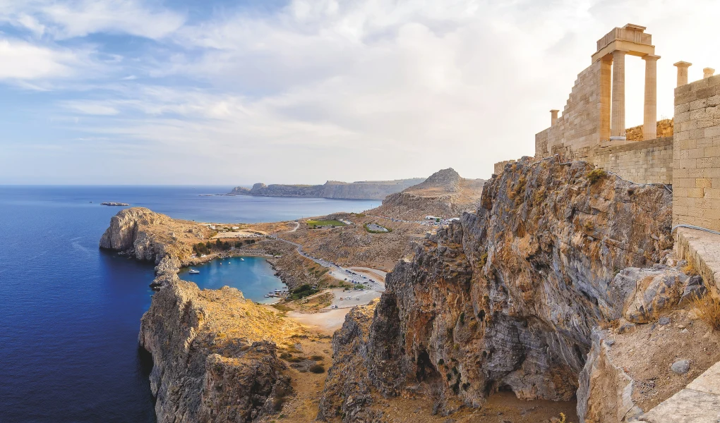 Widok antycznych ruin na stromym klifie nad brzegiem morza z malowniczą zatoką i rozległym, skalistym krajobrazem w tle.