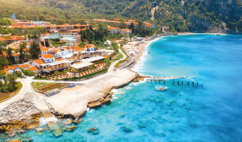 Widok na malowniczy kurort przy wybrzeżu, z jasnymi budynkami o pomarańczowych dachach, otoczonymi zielenią i górami oraz szeroką plażą schodzącą do turkusowego morza. W tle widać wzgórza porośnięte gęstym lasem.