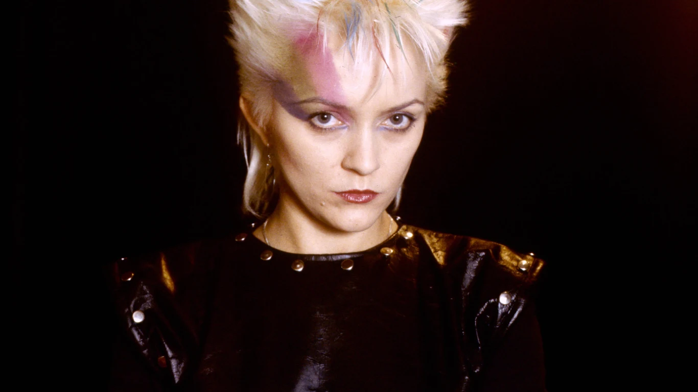 Małgorzata Ostrowska (1984 r.) Kobieta z jasnymi, krótkimi włosami ułożonymi w stylu punk, z intensywnym makijażem, ubrana w czarną, błyszczącą kurtkę ze zdobieniami i patrząca pewnie w stronę obiektywu, na ciemnym tle.