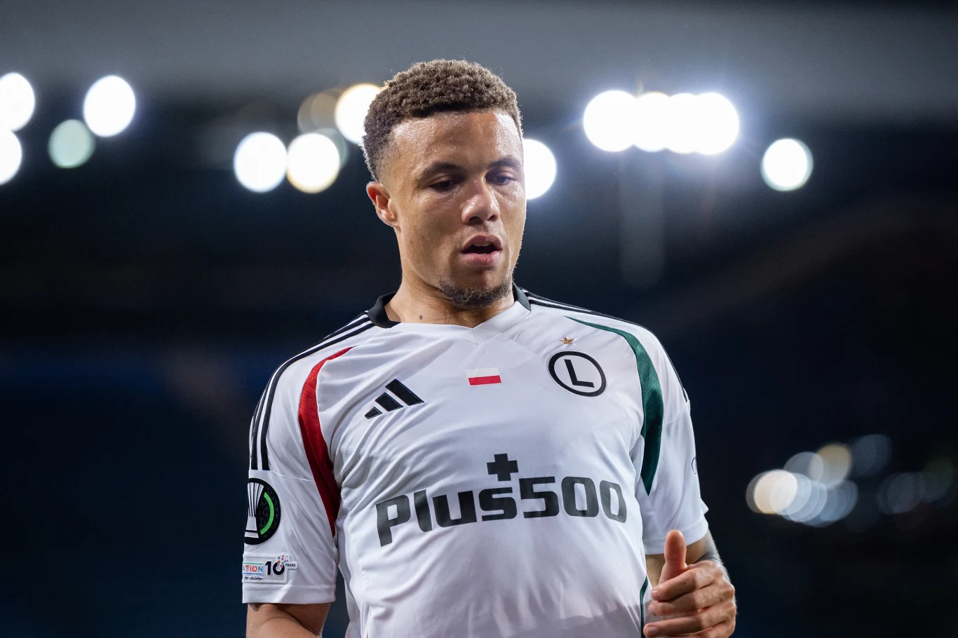 Piłkarz w białej koszulce klubu Legia Warszawa podczas meczu na stadionie, otoczony rozmytym tłem i światłami stadionowymi.