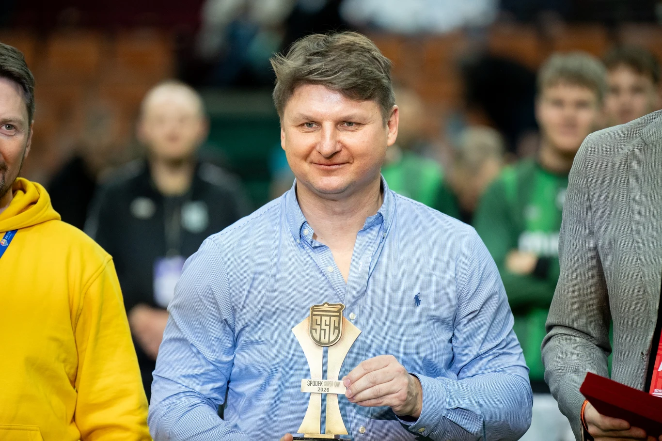 Mężczyzna w niebieskiej koszuli trzyma w rękach statuetkę z logo SSG, wokół niego inni uczestnicy wydarzenia, w tle osoby w sportowych strojach.