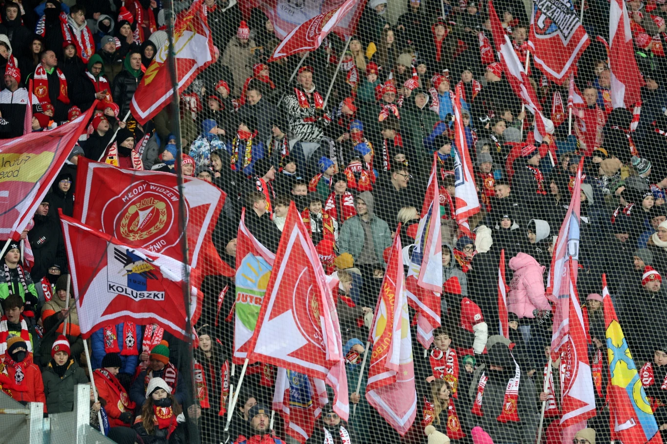 Tłum kibiców ubranych w czerwone barwy i szaliki klubowe, machających licznymi flagami klubowymi oraz innych drużyn, z widoczną siatką bezpieczeństwa oddzielającą trybuny od boiska.