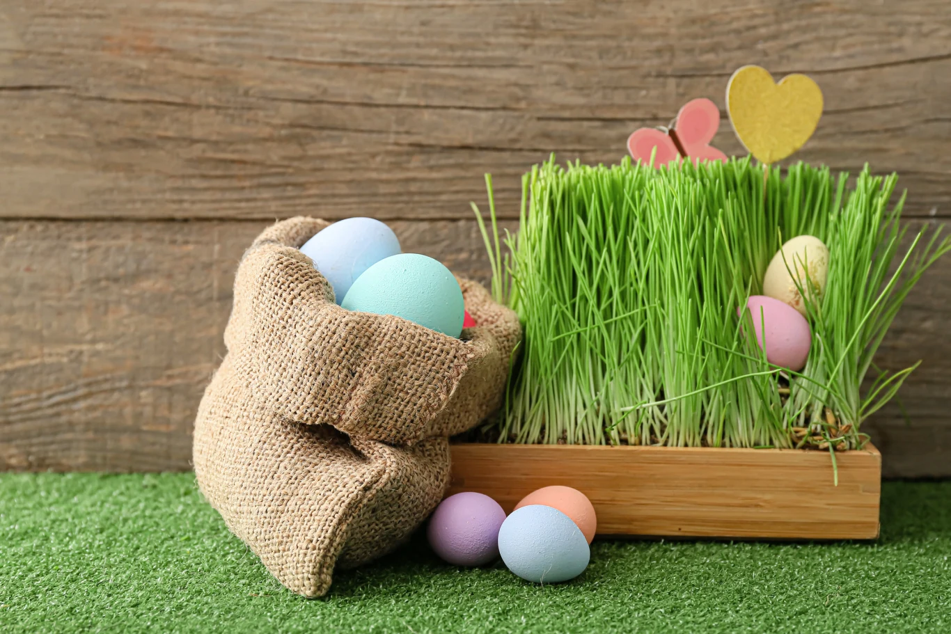 Kolorowe, pastelowe pisanki umieszczone w jutowym worku obok pojemnika z zieloną trawą ozdobioną papierowymi serduszkami i jajkami, wszystko na tle drewnianej ściany i zielonej sztucznej trawy.