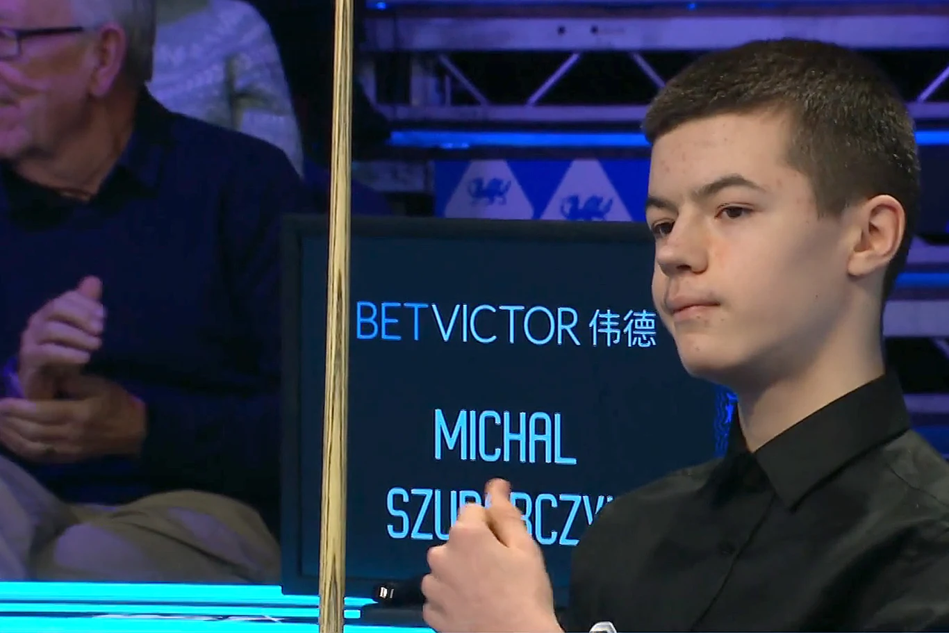 Młody mężczyzna ubrany na czarno trzyma kij bilardowy podczas turnieju snookera, w tle widoczna tablica z jego imieniem i nazwiskiem oraz logo sponsora.