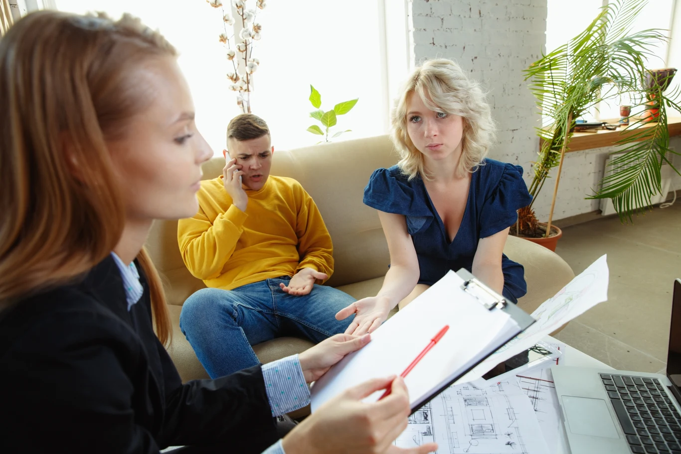 Ile kosztuje sporządzenie aktu notarialnego? Wielu kupujących nie bierze tego pod uwagę. Trzy osoby w biurze w trakcie rozmowy, kobieta z dokumentami i długopisem wyjaśnia coś parze siedzącej na kanapie. Mężczyzna w żółtym swetrze wygląda na zmartwionego i rozmawia przez telefon, kobieta obok niego słucha uważnie.