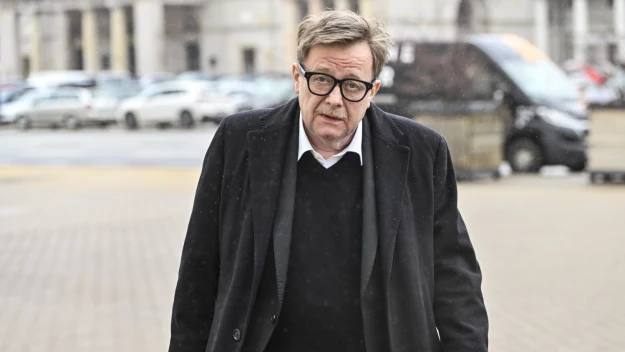 Mężczyzna w czarnym płaszczu i okularach idzie po chodniku miejskim, w tle zaparkowane samochody oraz budynki.