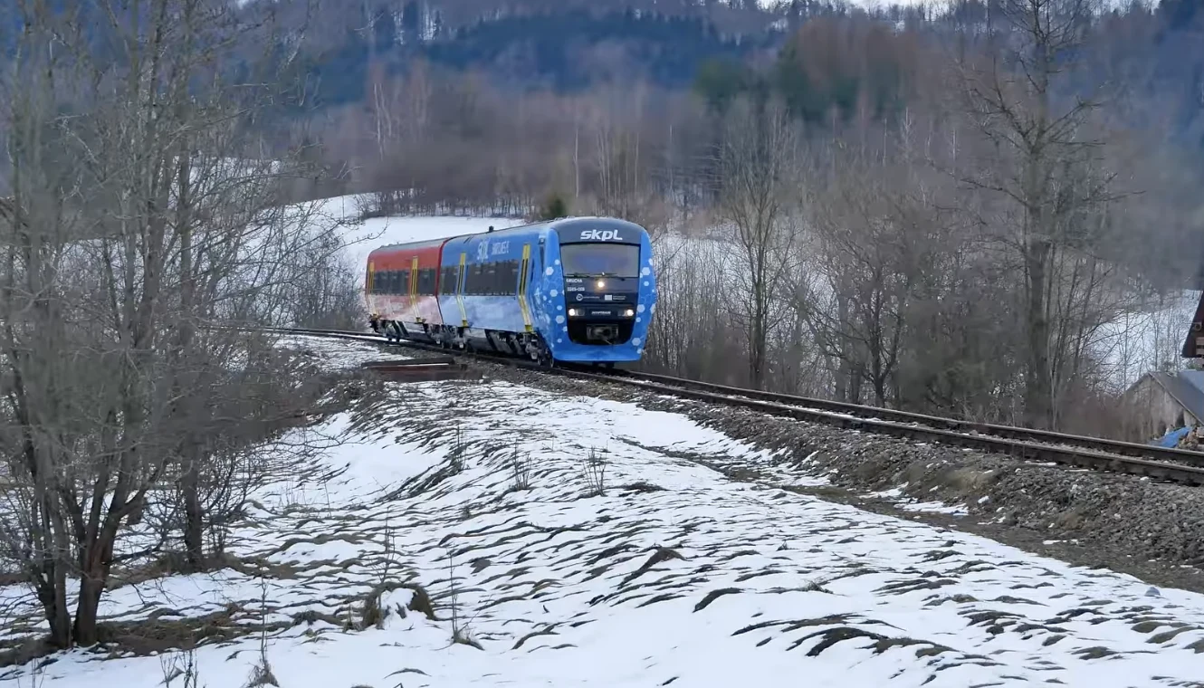 Pierwszy pociąg wodorowy SKPL Cargo wyjechał na tory w Polsce. Nowoczesny, niebiesko-czerwony pociąg poruszający się po zakręcie torów kolejowych przez zimowy krajobraz z lekkim śniegiem i bezlistnymi drzewami w tle.