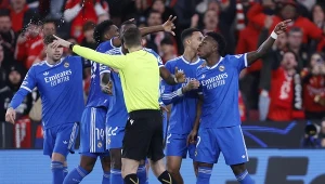 Piłkarze drużyny w niebieskich strojach dyskutują z sędzią podczas meczu piłkarskiego, tłum kibiców widoczny w tle.