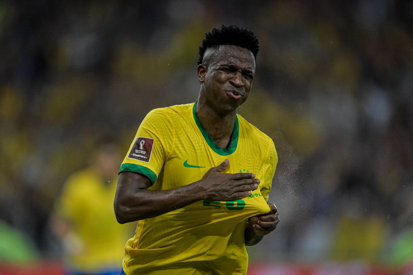 Piłkarz w żółtej koszulce reprezentacji Brazylii ściska swoją koszulkę na piersi, wyrażając silne emocje podczas meczu, tło stanowią rozmyci zawodnicy i trybuny.