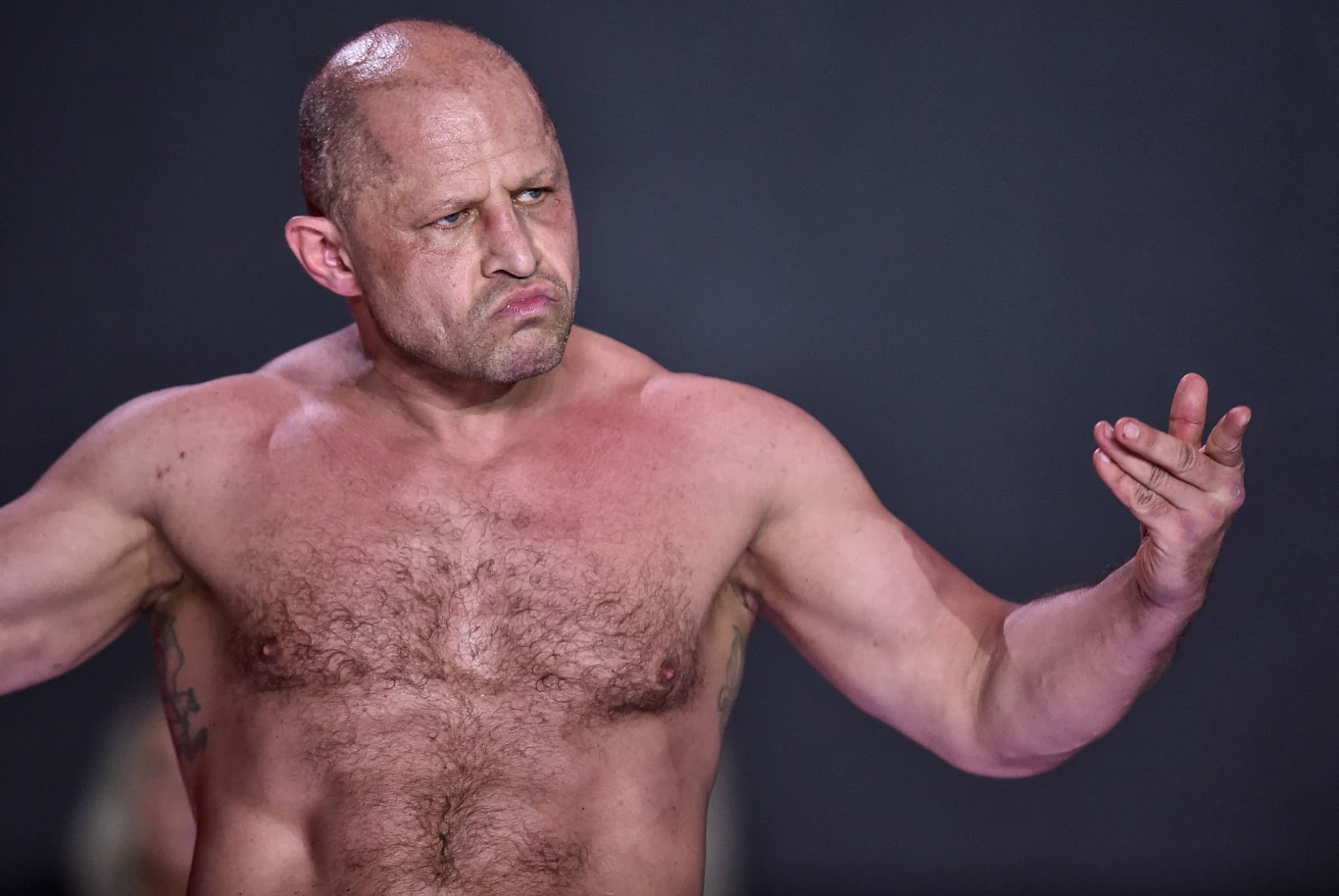 Jacek Murański Umięśniony, łysy mężczyzna z wyrazistą miną stoi z odkrytym torsem na ciemnym tle, jego ręce są wyciągnięte, a palce wskazują gest sugerujący wyzwanie lub pewność siebie.