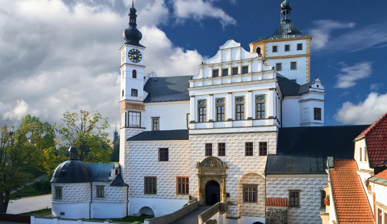 Co warto zobaczyć w zamku Pardubice? Renesansowy zamek z charakterystycznymi wieżami, białą elewacją i zabytkowymi oknami, otoczony drzewami oraz częścią brukowanego dziedzińca pod błękitnym niebem z chmurami.