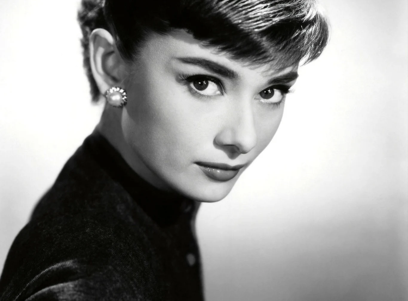 Audrey Hepburn Portret młodej kobiety o ciemnych włosach, z eleganckimi perłowymi kolczykami oraz wyrazistym spojrzeniu, wykonany w stylu czarno-białym, podkreślający subtelność i delikatność rysów twarzy.