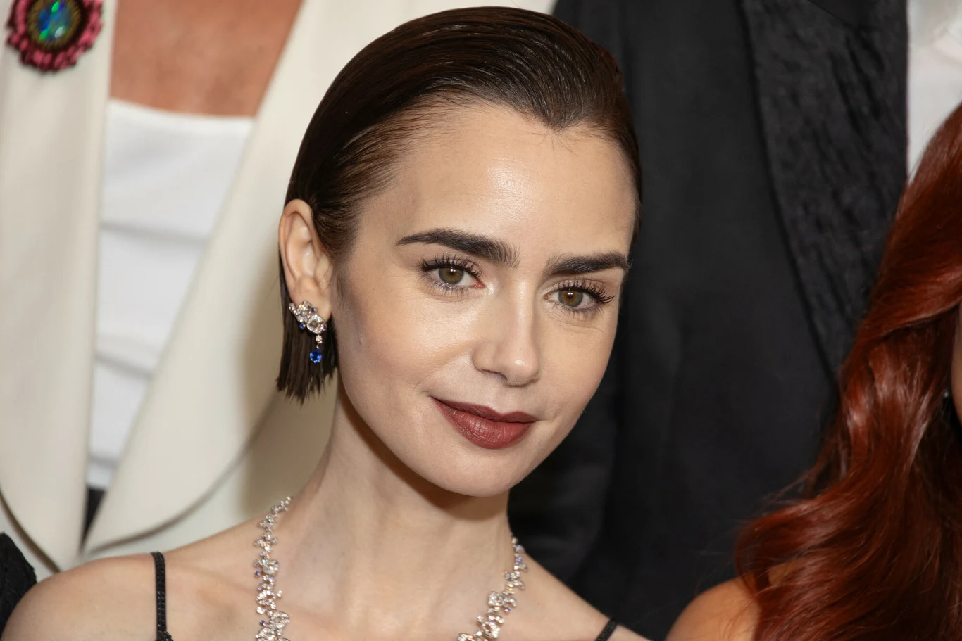 Lily Collins Młoda kobieta o ciemnych, wygładzonych do tyłu włosach z delikatnym makijażem, uśmiecha się subtelnie, mając na sobie ozdobne kolczyki, elegancki srebrny naszyjnik oraz czarną, cienką sukienkę. W tle widoczne fragmenty stroju innych osób, w tym białej ...