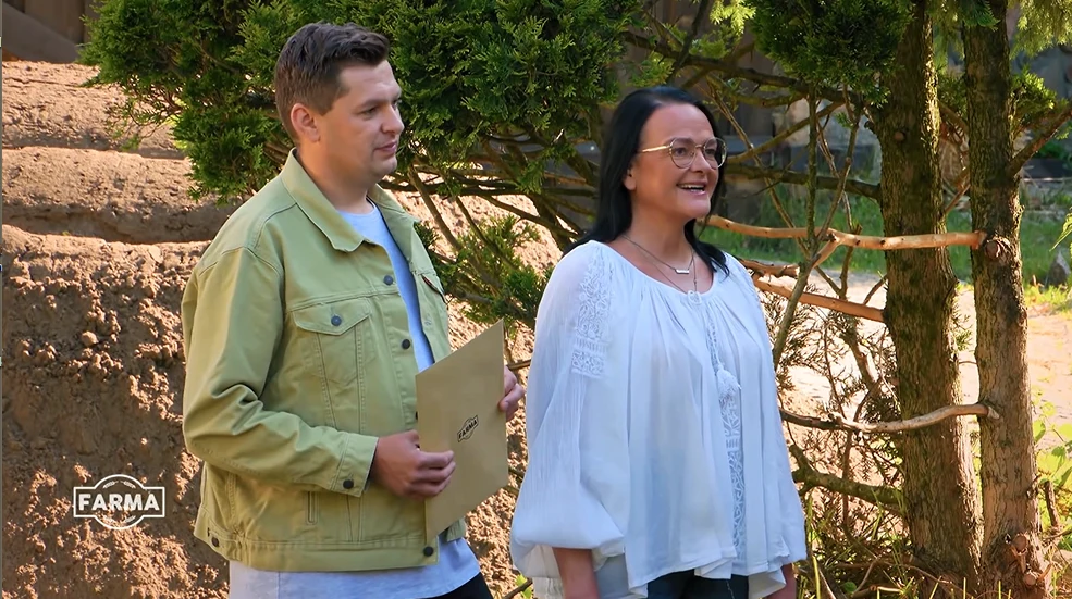 Farmerów odwiedzili Szymon i Basia Dwie osoby stoją obok siebie na tle zieleni, jedna z nich trzyma kopertę, obie uśmiechnięte i zwrócone lekko w prawo; z tyłu drzewa i ogrodzenie z gałęzi.